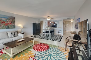 A modern living room with a sofa, coffee table, and a colorful rug. There is a dining area with chairs and a table, and a kitchen in the background.