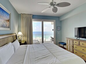Et hotelværelse med seng, ventilator, fjernsyn, kommode, stol og balkon med havudsigt.