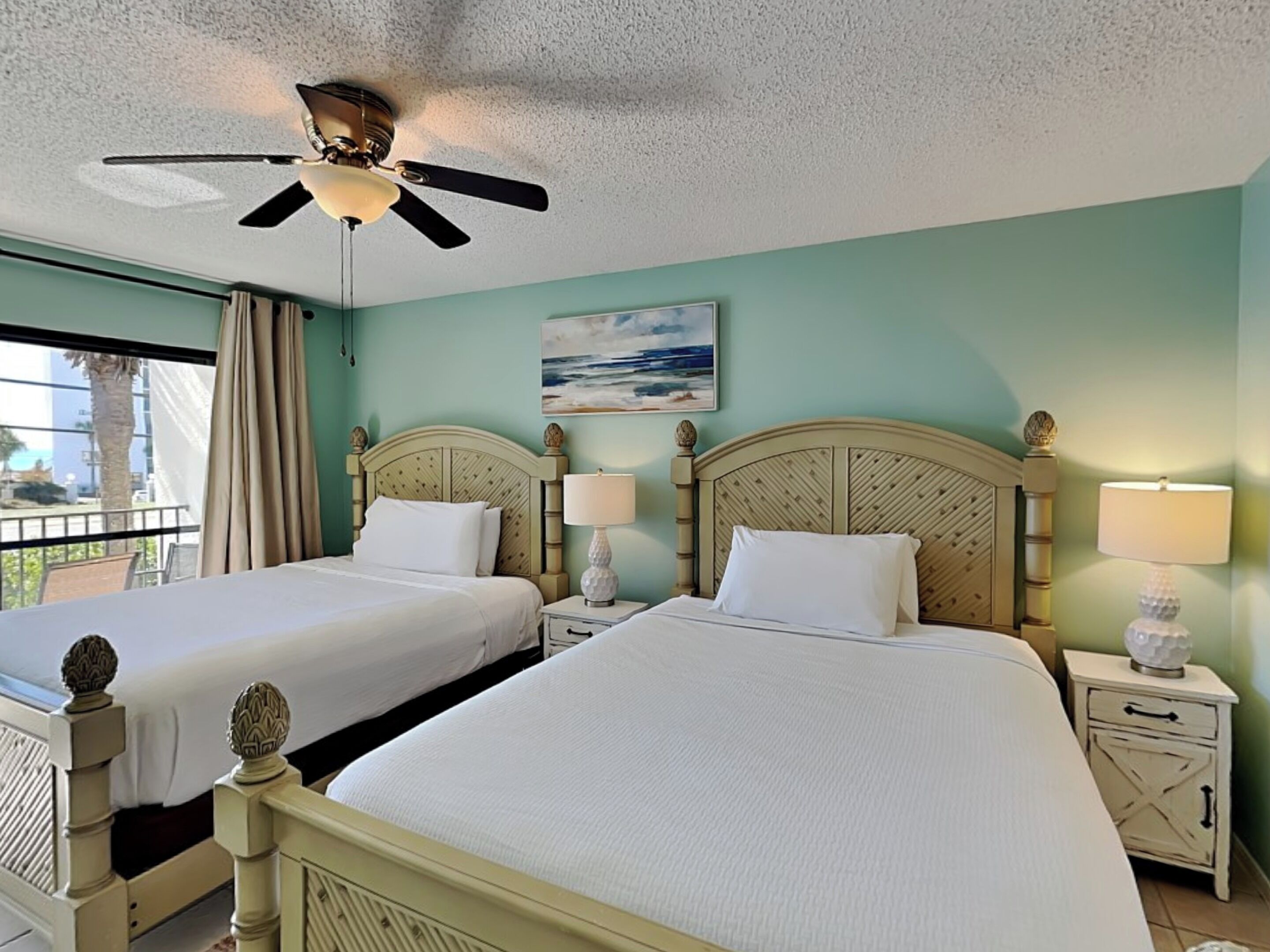 A hotel room with two beds, a ceiling fan, a painting of the ocean, and a balcony with a view of palm trees.