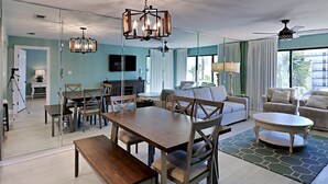 A spacious living area with a dining table, sofas, and a coffee table. There is a large window providing natural light and a view of the outdoors. - Edgewater Villa #3315 | Beach, Golf, & 11 Pools! (Panama City Beach)