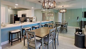 A modern kitchen with white cabinets, a central island, and a dining area with a wooden table and chairs. - Edgewater Villa #3315 | Beach, Golf, & 11 Pools! (Panama City Beach)