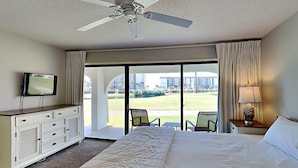Habitación de hotel con una cama grande, ventilador de techo, televisión, vistas al exterior y un escritorio con lámpara.
