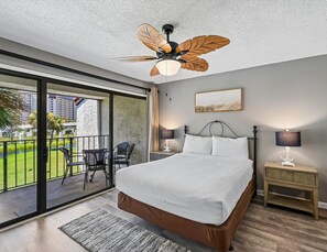 Un dormitorio con cama, mesita de noche, lámpara, ventilador de techo, vista a un balcón y un cuadro en la pared.