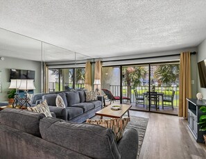 A modern living room with a grey sofa, a coffee table, and a balcony with outdoor furniture. - Edgewater Villa #512 | Golf, Beach Access, Tennis, & More! (Panama City Beach)
