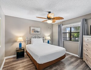Une chambre à coucher comprenant un lit, deux tables de chevet avec des lampes, un ventilateur de plafond, une fenêtre avec des rideaux et une commode.