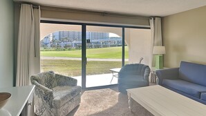 Ein Hotelzimmer mit einer großen Schiebeglasdoor zum Balkon mit Blick auf einen Golfplatz und Gebäude. Es gibt ein blaues Sofa, einen gemusterten Sessel und einen kleinen Tisch mit einer Lampe.