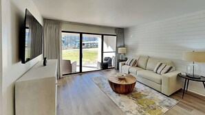 A modern living room with a sofa, a coffee table, a TV mounted on the wall, and a large sliding glass door leading to an outdoor area. - Edgewater Villa #505 | 11 Pools, Golf Course, & More (Panama City Beach)