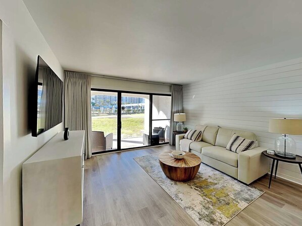 A modern living room with a sofa, a coffee table, a TV mounted on the wall, and a large sliding glass door leading to an outdoor area. - Edgewater Villa #505 | 11 Pools, Golf Course, & More (Panama City Beach)