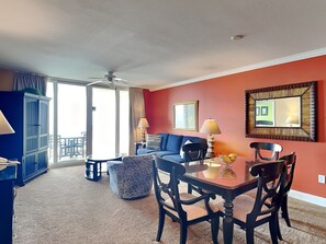 A hotel room with a blue sofa, a round coffee table, a dining table with chairs, a blue cabinet, and a large window with curtains. - Emerald Beach #1825 | Dining, Pool, Gym, & More! (Panama City Beach)