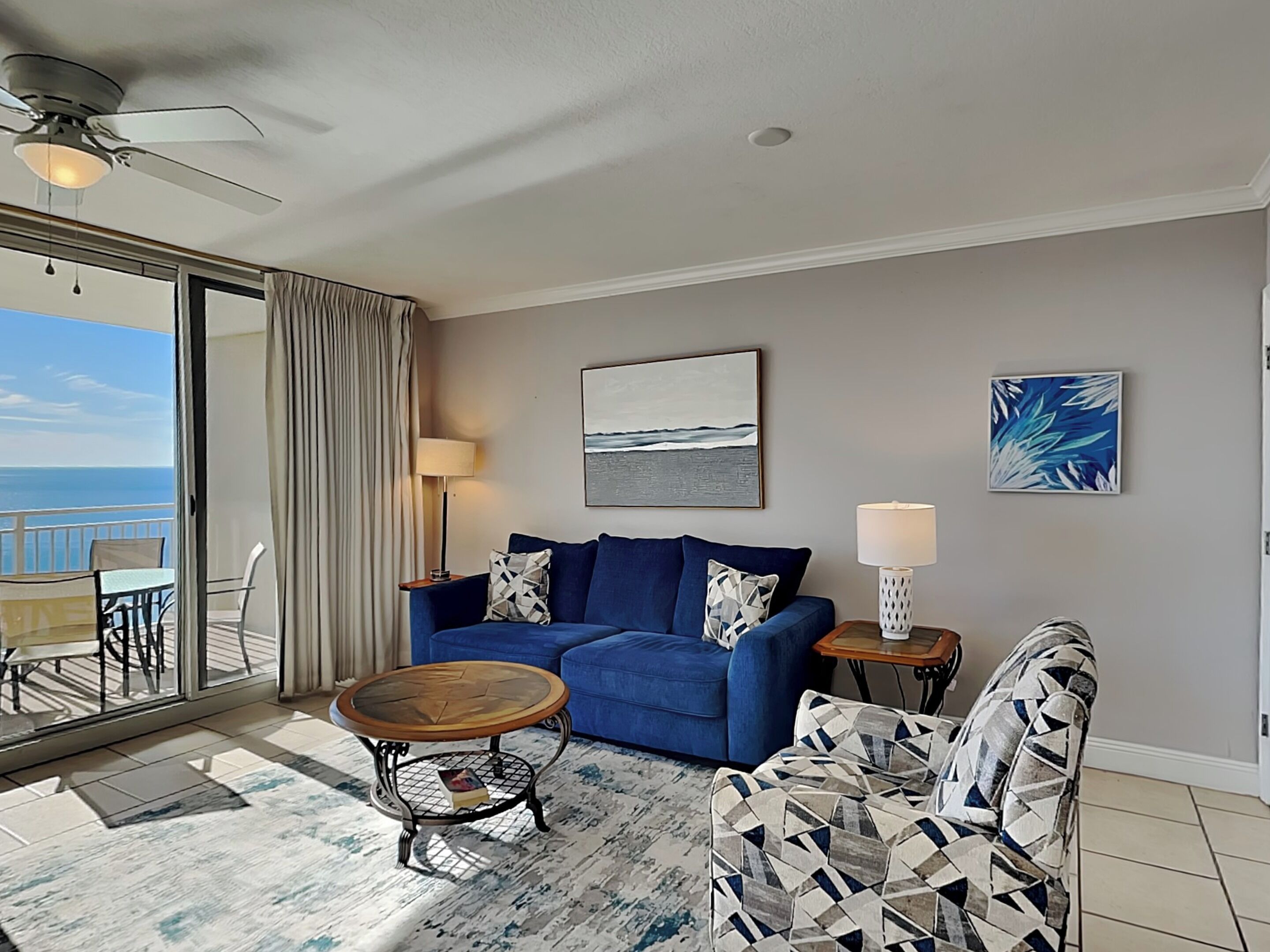 Una sala de estar moderna con un sofá azul, un sillón estampado, una mesita de centro y vistas al océano a través de una puerta corrediza.