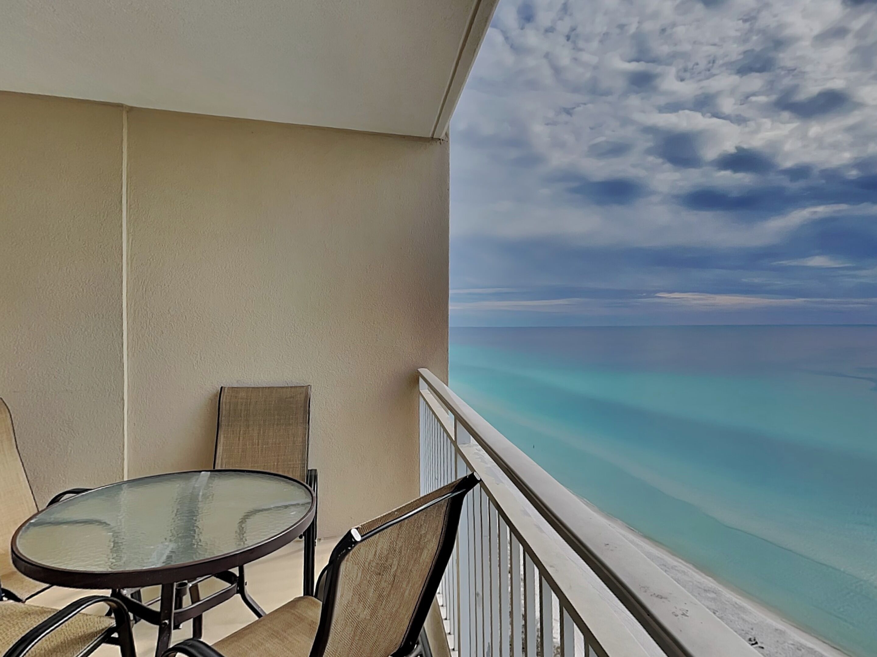 Ein Balkon mit Glastisch und Stühlen, der Blick auf ein klares türkisfarbenes Meer und einen bewölkten Himmel bietet.