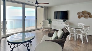 Un balcon spacieux avec une porte vitrée donnant sur une piscine, un coin repas avec une table ronde et des chaises, un canapé, un téléviseur et des œuvres d’art décoratives sur les murs.