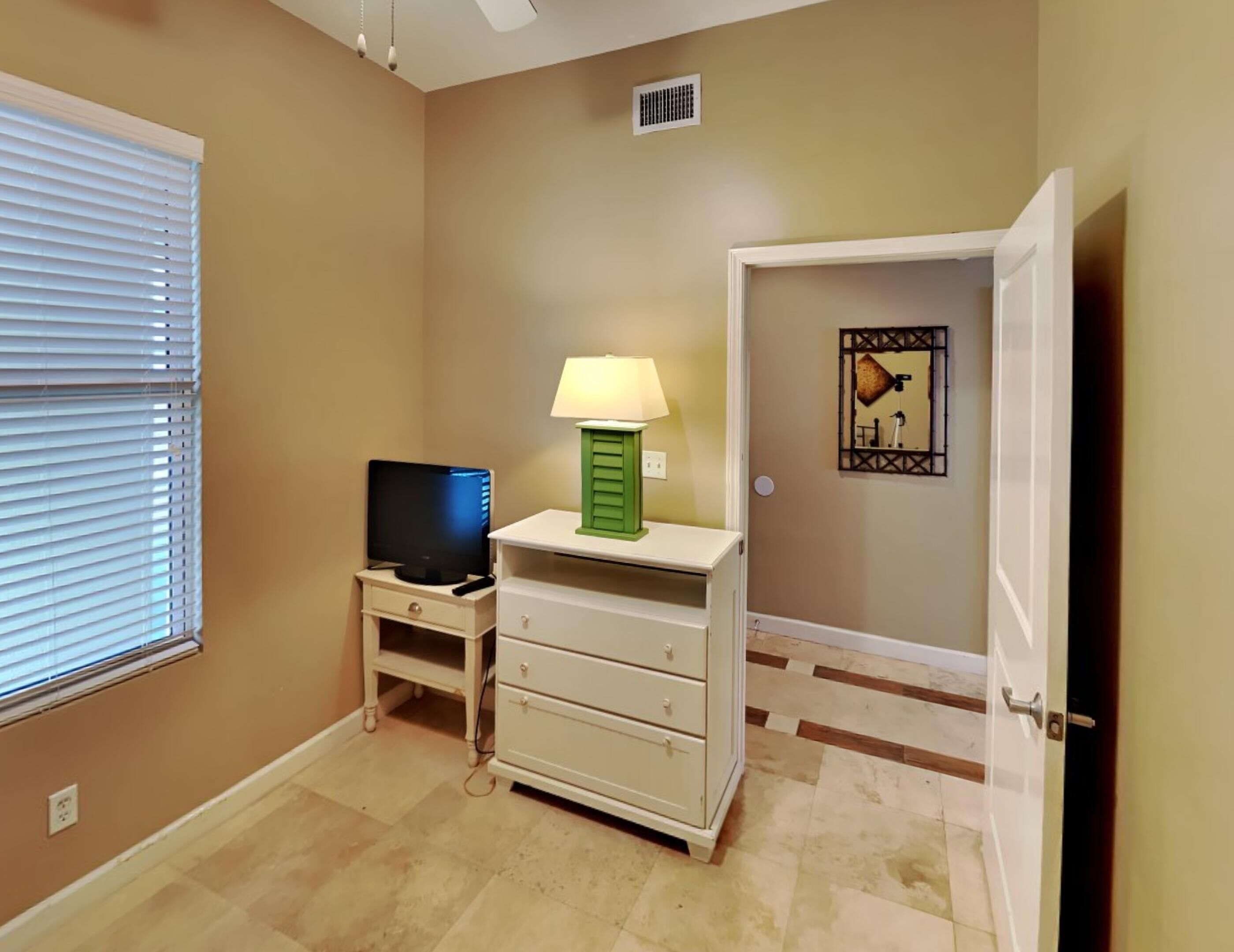 A room with beige walls, a television on a white stand, a green lamp, a white dresser, and a door with a framed picture.