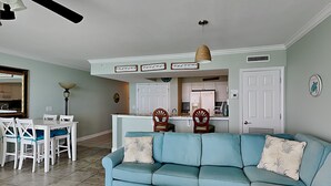 A living room with a blue sofa, white chairs, a dining table, and a kitchen area in the background.