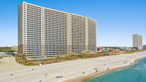 A large hotel with multiple floors and balconies, situated next to a beach with clear blue water and sandy shores.