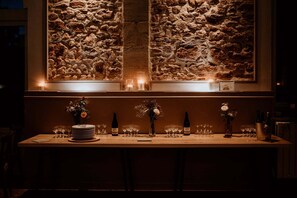 A bar area with a stone wall backdrop, candles, and a neatly arranged table with wine bottles, glasses, and a vase of flowers.