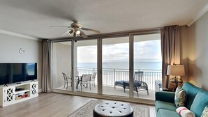 Ein großzügiges Wohnzimmer mit großem Fernseher, Deckenventilator, einer Couch und einem Balkon mit Meerblick.