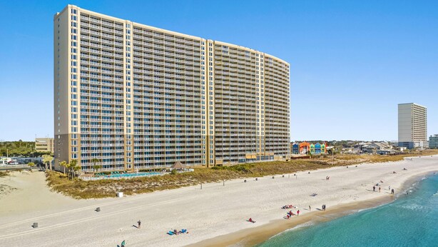 Een groot hotel met meerdere verdiepingen en balkons, gelegen naast een strand met helderblauw water en zandstranden.