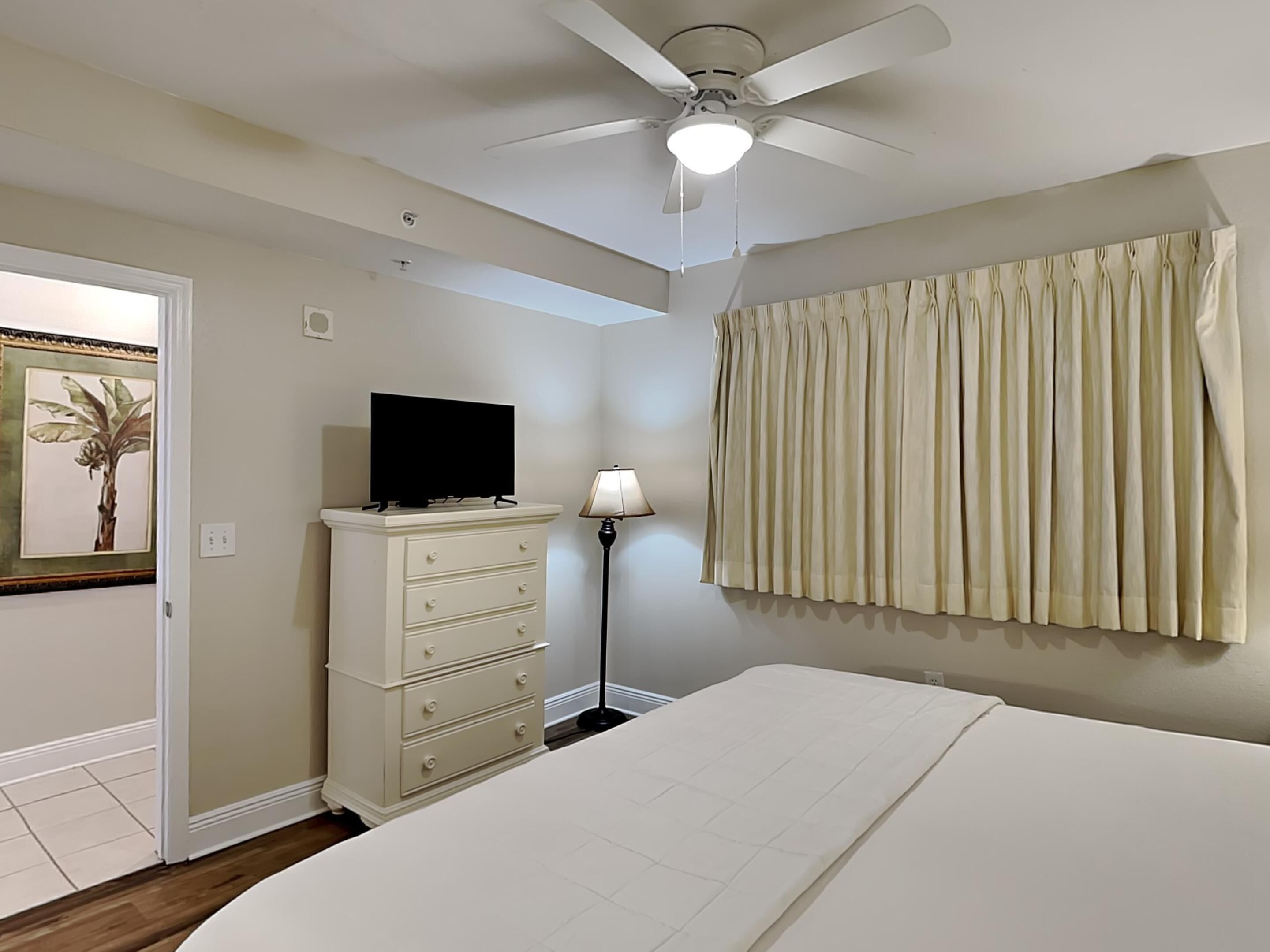 Un dormitorio con cama, cómoda, ventilador de techo, lámpara y ventana con cortinas.