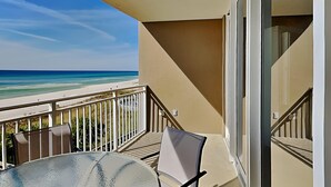 A balcony with a glass table and chairs, overlooking a beach and ocean.