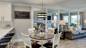A modern kitchen with white cabinetry, a dining area with a round table and white chairs, and a living room with a grey sofa and lamps. - Edgewater Villa #909| Full-service resort! (Panama City Beach)