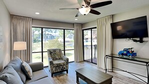 A spacious living room with a ceiling fan, a sofa, a patterned armchair, a coffee table, a TV mounted on the wall, and a view of the outdoors through large windows. - Edgewater Villa #708 | Full-service resort! (Panama City Beach)