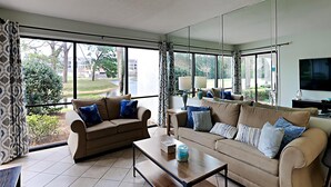 Amplia sala de estar con dos sofás, una mesa de centro y un televisor. Ventanas grandes ofrecen vistas al exterior.