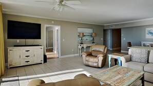 A spacious living room with a ceiling fan, a flat-screen TV, a leather armchair, a coffee table, and a sofa. - Emerald Beach #2435 | Stunning Views + Resort Amenities! (Panama City Beach)