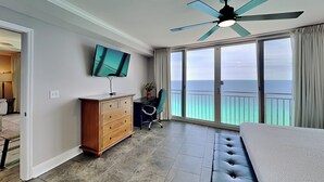 Ein Hotelzimmer mit einer großen Schiebeglastür zum Meer, einem Deckenventilator, einem Flachbildfernseher, einer Holzkommode und einer gepolsterten Bank.