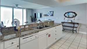 A kitchen with granite countertops, white cabinets, a stainless steel sink, and a dining area with a table and chairs. - Emerald Beach #328 | Views, Pool, Dining! (Panama City Beach)
