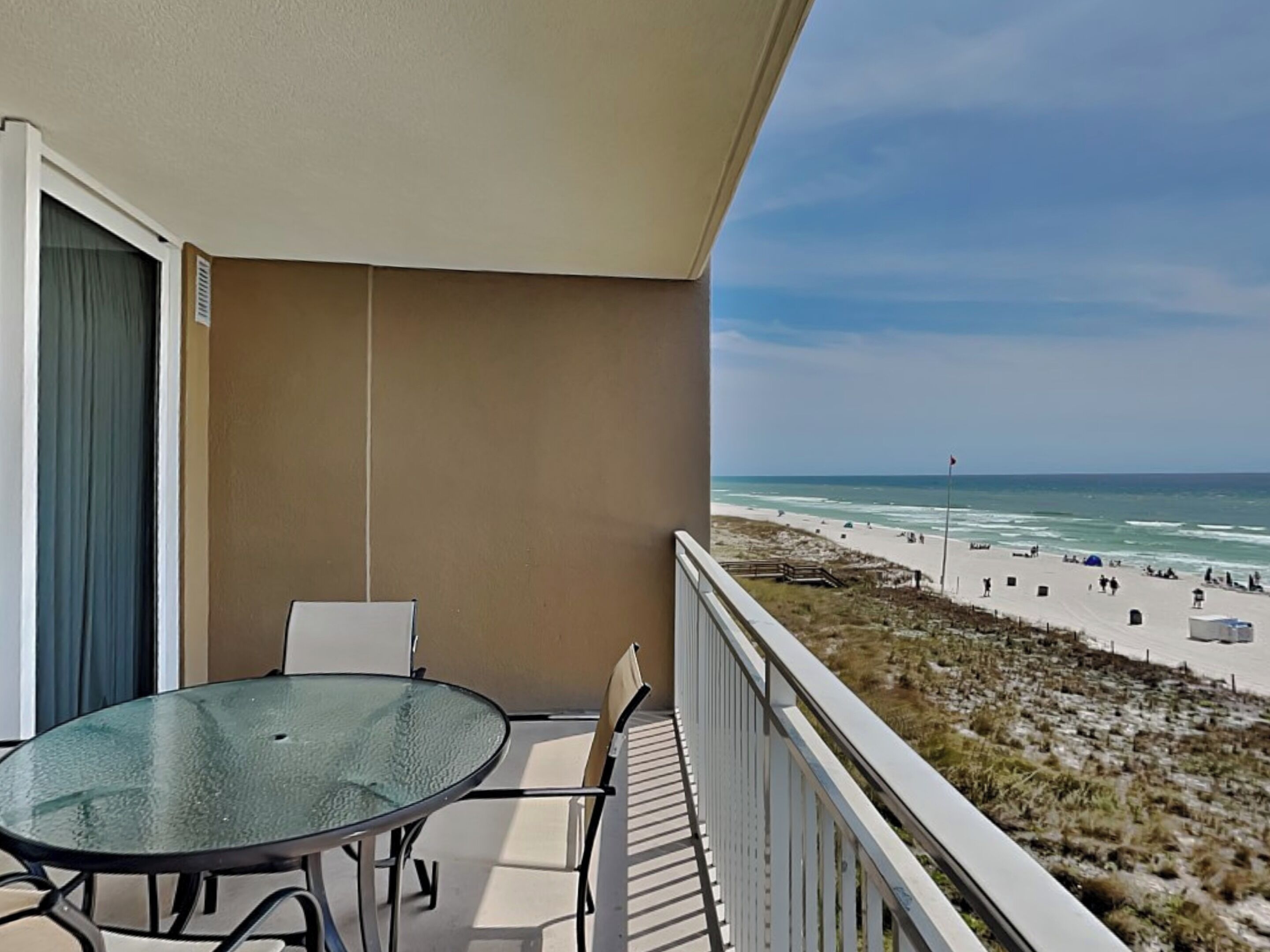 A balcony with a glass table and chairs, overlooking a beach with people and a flagpole.