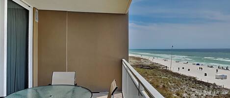 A balcony with a glass table and chairs, overlooking a beach with people and a flagpole.