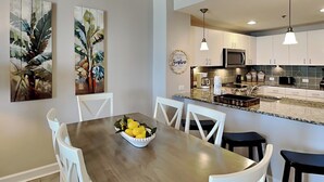 A modern kitchen with white cabinets, a granite countertop, and a built-in microwave. There is a dining area with a wooden table and white chairs, and a bowl of lemons on the table. - Grand Panama Beach Resort - The Grand #1304 (Panama City Beach)