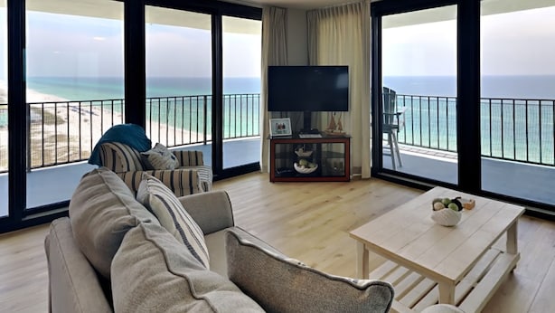 A living room with a sofa, a coffee table, and a TV. The room has large windows with a view of the beach and ocean. - Watercrest #1401 | Warm turquoise waters! (Panama City Beach)