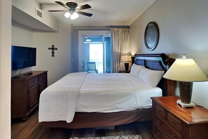 A bedroom with a bed, a ceiling fan, a TV, a dresser, a lamp, and a view of the balcony. - The Grand #403 | Hot Tubs, Tiki Bar, & More! (Panama City Beach)