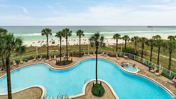 A swimming pool with a beach view, palm trees, and a pier in the background.