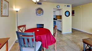 A dining area with a red table, wooden chairs, a ceiling fan, and a kitchen in the background. - Sunbird Beach Resort #204 East | Gulf Front (Panama City Beach)