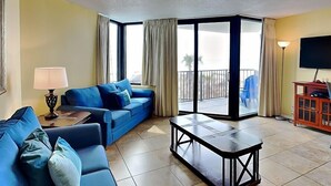A living room with a blue sofa, a coffee table, a lamp, and a TV. - Sunbird Beach Resort #204 East | Gulf Front (Panama City Beach)