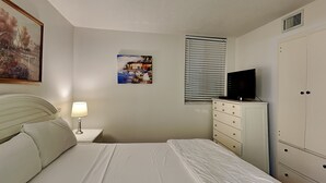 A bedroom with a bed, a nightstand with a lamp, a dresser, a television, and a painting on the wall. - Sunbird Beach Resort #204 East | Gulf Front (Panama City Beach)