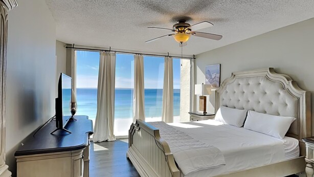 A bedroom with a large bed, a ceiling fan, a TV, and a view of the ocean. - Long Beach I #503 | Balcony Views! (Panama City Beach)