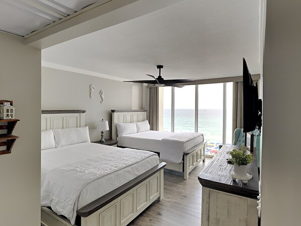 Habitación de hotel con dos camas, ventilador de techo, televisión y vista al océano.