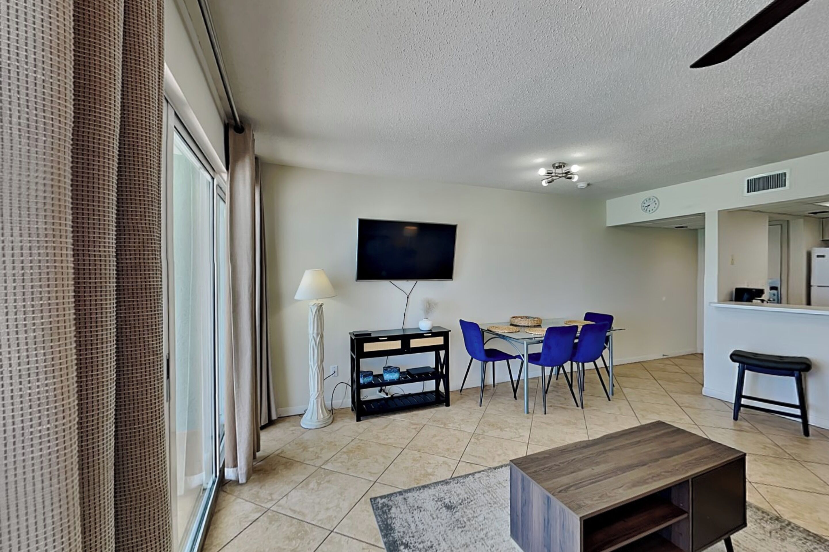Una sala de estar moderna con televisor de pantalla plana, un comedor con sillas azules, una mesa de centro y una cocina visible a través de una puerta abierta.