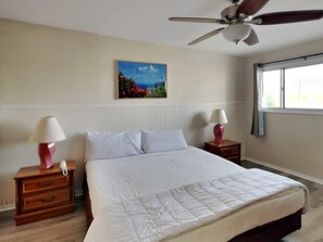 Een slaapkamer met een bed, twee nachtkastjes met lampen, een plafondventilator, een raam met gordijnen en een schilderij aan de muur.