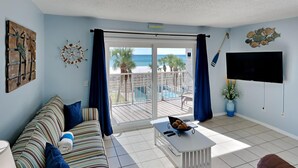 Un salon avec un canapé, une table basse et un téléviseur. Il y a un balcon avec vue sur la plage.