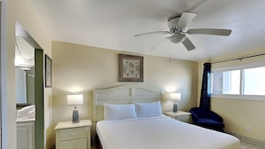 Une chambre d’hôtel avec un grand lit, deux tables de chevet avec des lampes, un ventilateur de plafond, une fenêtre avec des rideaux et un tableau encadré au mur.