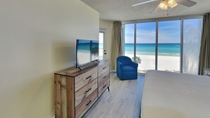 Ein Schlafzimmer mit großem Fenster und Blick auf den Strand, Deckenventilator und einem Holzkasten.
