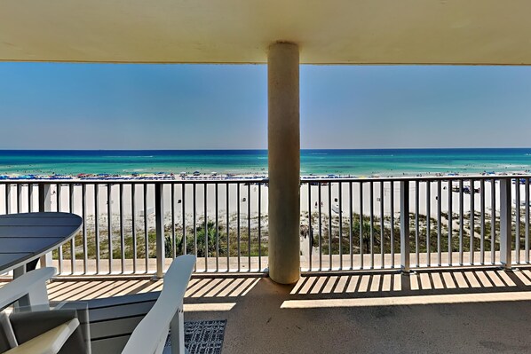 A balcony with a view of a beach, featuring a table and chairs, a railing, and a concrete pillar.