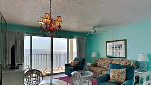 A living room with a view of the ocean, a chandelier, teal sofas, and a ceiling fan. - Long Beach I #702 | Gulf-Front Views! (Panama City Beach)