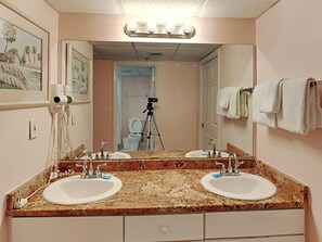 A bathroom with two sinks, a large mirror, and a hairdryer on the wall. - Long Beach I #702 | Gulf-Front Views! (Panama City Beach)