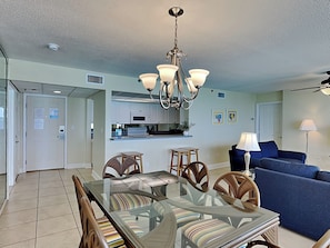 A spacious living area with a glass dining table surrounded by wicker chairs, a blue sofa, and a modern kitchen in the background.